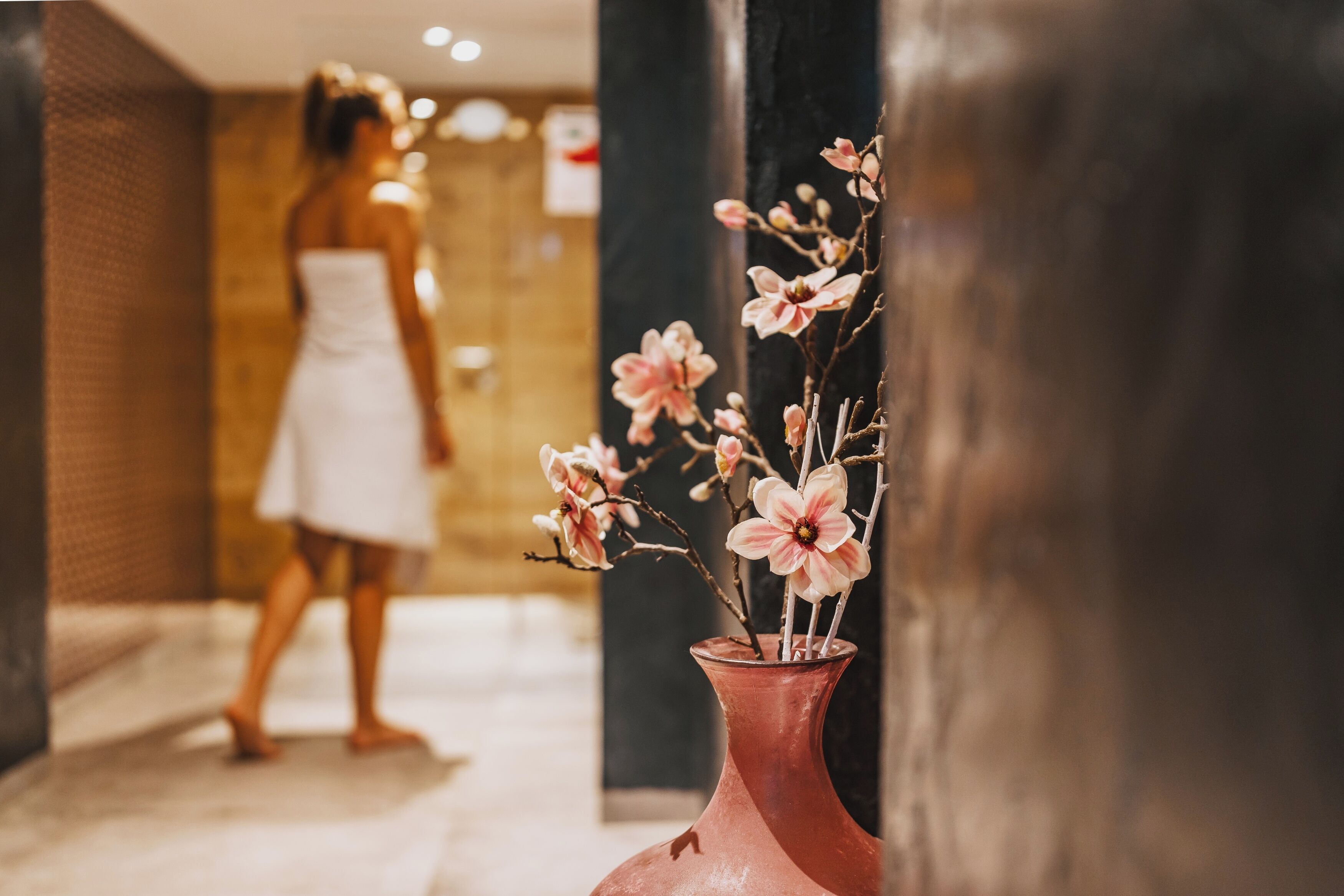 Un couloir orné d’un vase de fleurs roses, une personne vêtue d’une robe blanche et un pilier noir.