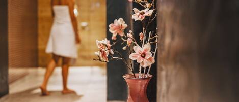 Un couloir orné d’un vase de fleurs roses, une personne vêtue d’une robe blanche et un pilier noir.