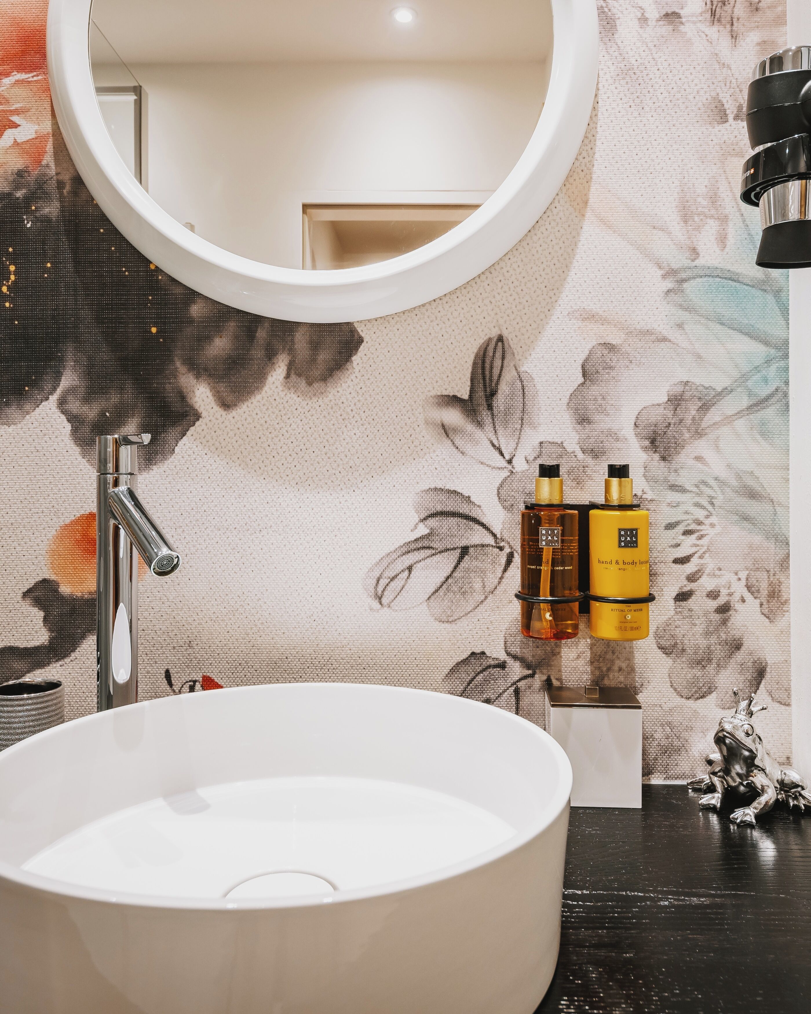 A bathroom with a round mirror, a white sink, and a floral-patterned wall.