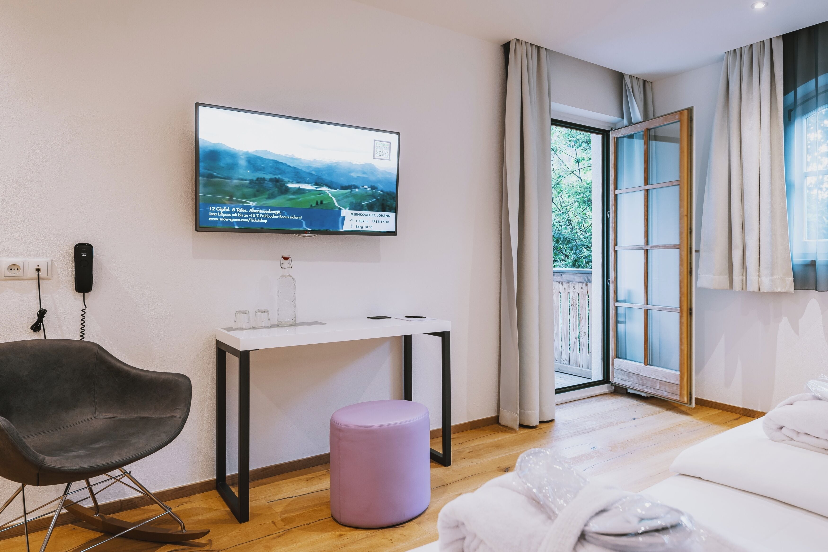 Ein modernes Hotelzimmer mit großem Bildschirm an der Wand, der eine schöne Landschaft zeigt, einem kleinen Schreibtisch mit Stuhl, einem großen Fenster mit Vorhängen und einem ordentlich bezogenen Bett.