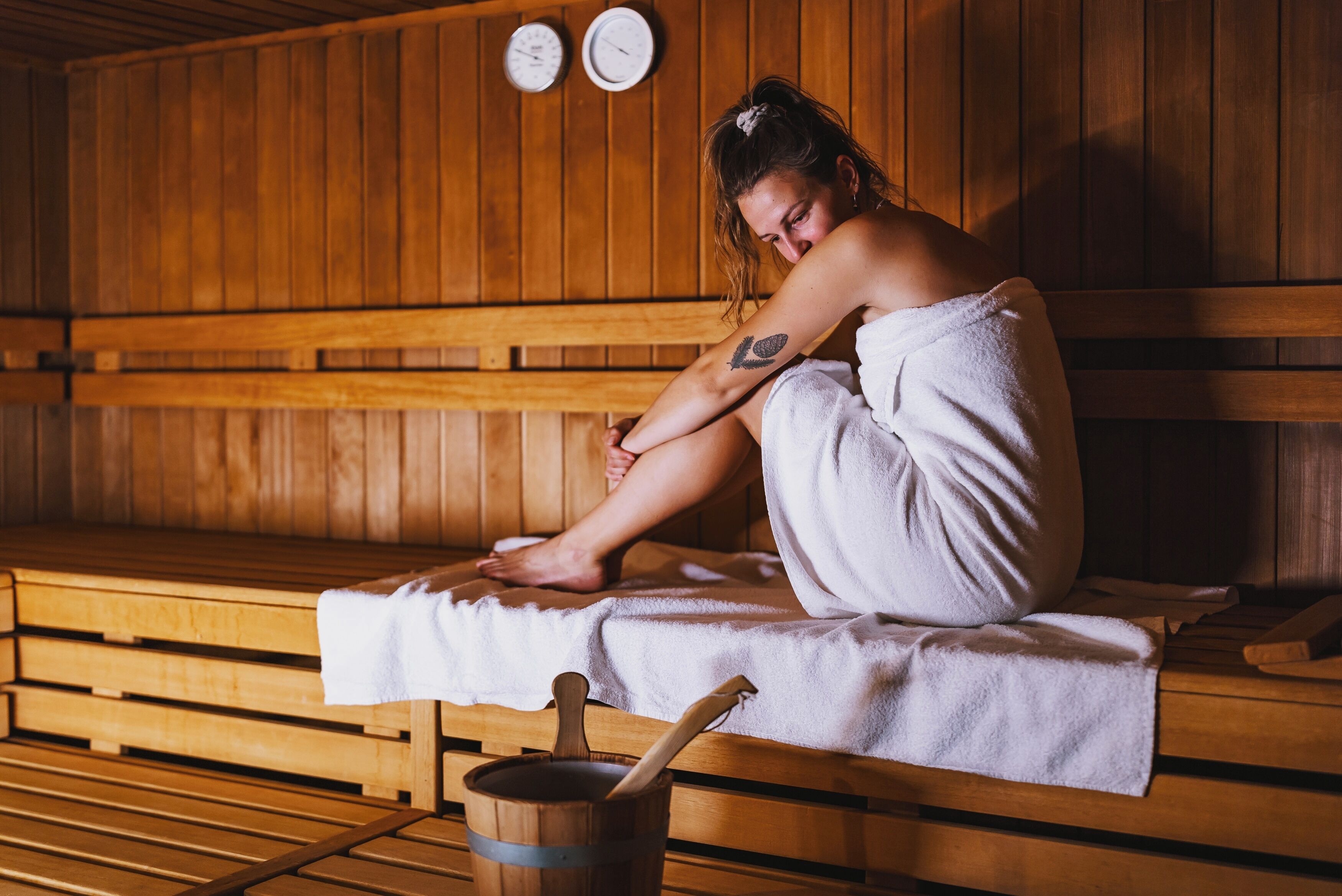 A person sitting on a wooden bench in a sauna room with a towel wrapped around them, wooden benches, and a wooden bucket with a stick.