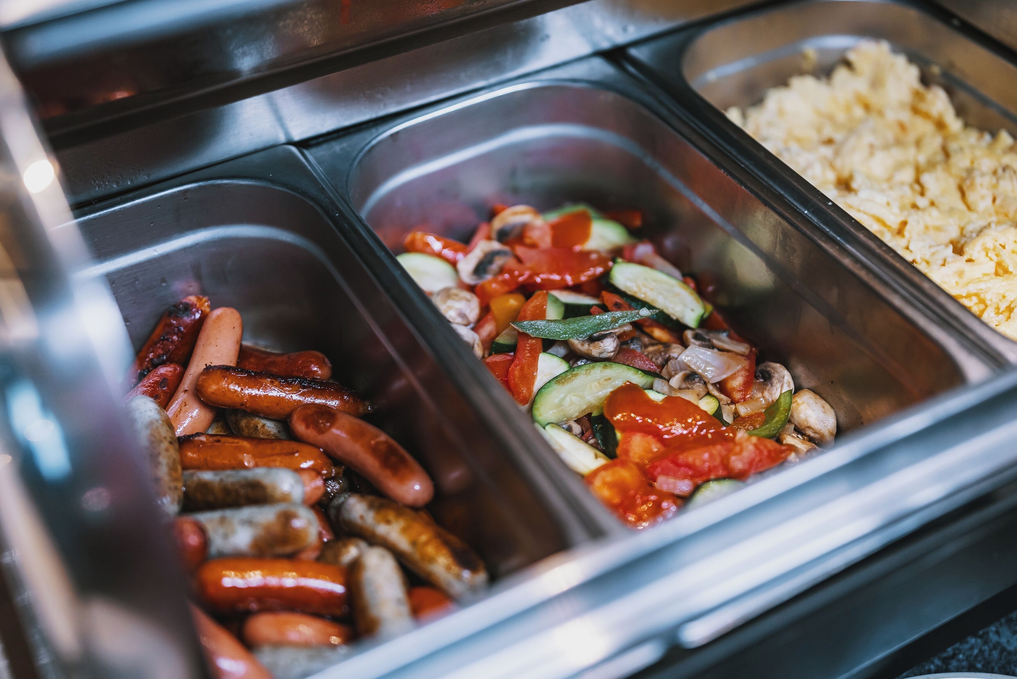 A buffet with various food options including sausages, vegetables, and scrambled eggs.