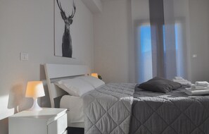 A neatly made bed with a quilted cover, a bedside lamp, a wall-mounted artwork of a deer, and a window with curtains.