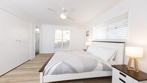 A bedroom with a bed, bedside lamps, a ceiling fan, and a window with blinds. - Tiro Moana beautifully renovated pet friendly home (Forster)