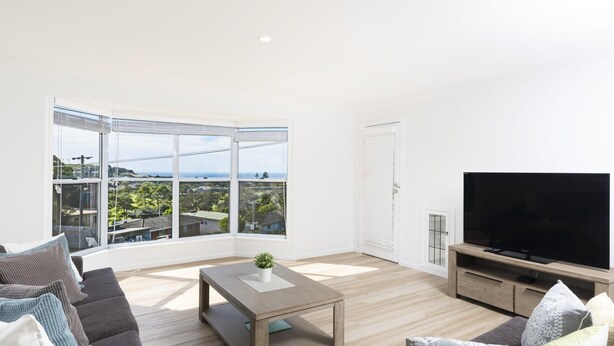 A modern living room with a large TV, a sofa, a coffee table, and a view of a coastal area. - Tiro Moana beautifully renovated pet friendly home (Forster)