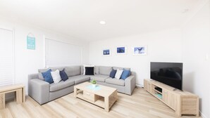 A modern living room with a grey sectional sofa, a wooden coffee table, a TV on a wooden stand, and a wall with three framed pictures. - Tiro Moana beautifully renovated pet friendly home (Forster)
