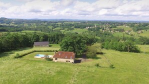 Een landelijk landschap met een huis, voorzien van een zwembad, omringd door groene velden en bomen.