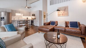 A modern living room with a brown leather sofa, a white armchair, and a coffee table. A kitchen is visible in the background with stainless steel appliances.