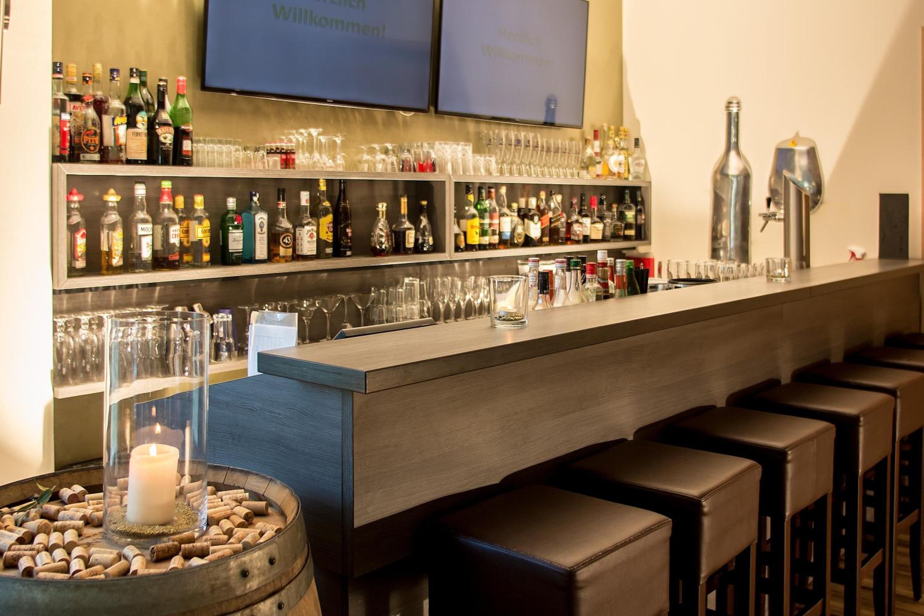Una zona de bar con estanterías llenas de botellas de licor, vasos y taburetes.
