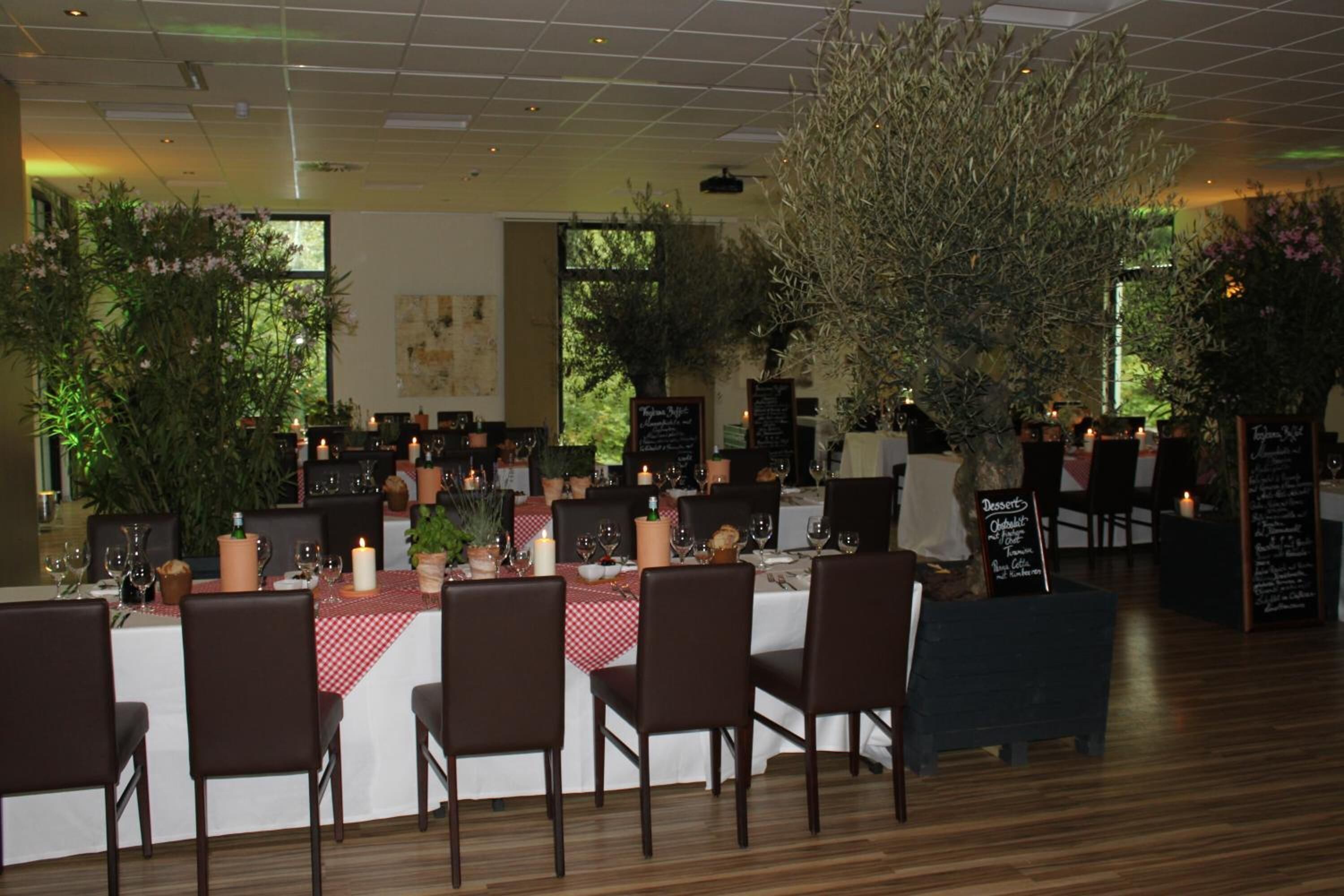 Un restaurant avec de nombreuses tables dressées pour le service, entouré de plantes en pot et d’un grand arbre.