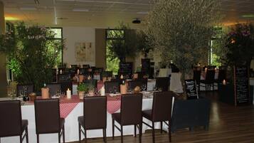 Un restaurant avec de nombreuses tables dressées pour le service, entouré de plantes en pot et d’un grand arbre.