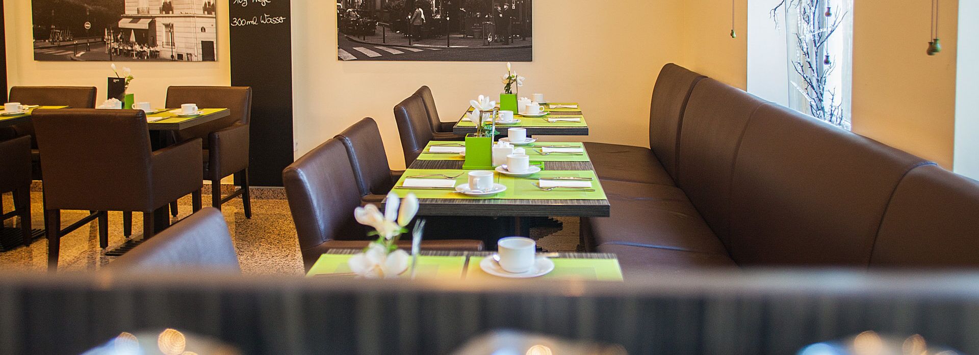 A restaurant with booths and tables, some with green placemats. There are framed pictures on the walls and a window with a view.