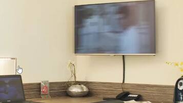 A hotel reception area with a flat-screen TV mounted on the wall, a laptop on the desk, a vase with flowers, a phone, and a coffee machine.