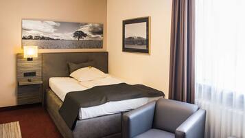 A hotel room with a bed, a bedside table with a lamp, a chair, and two framed pictures on the wall.