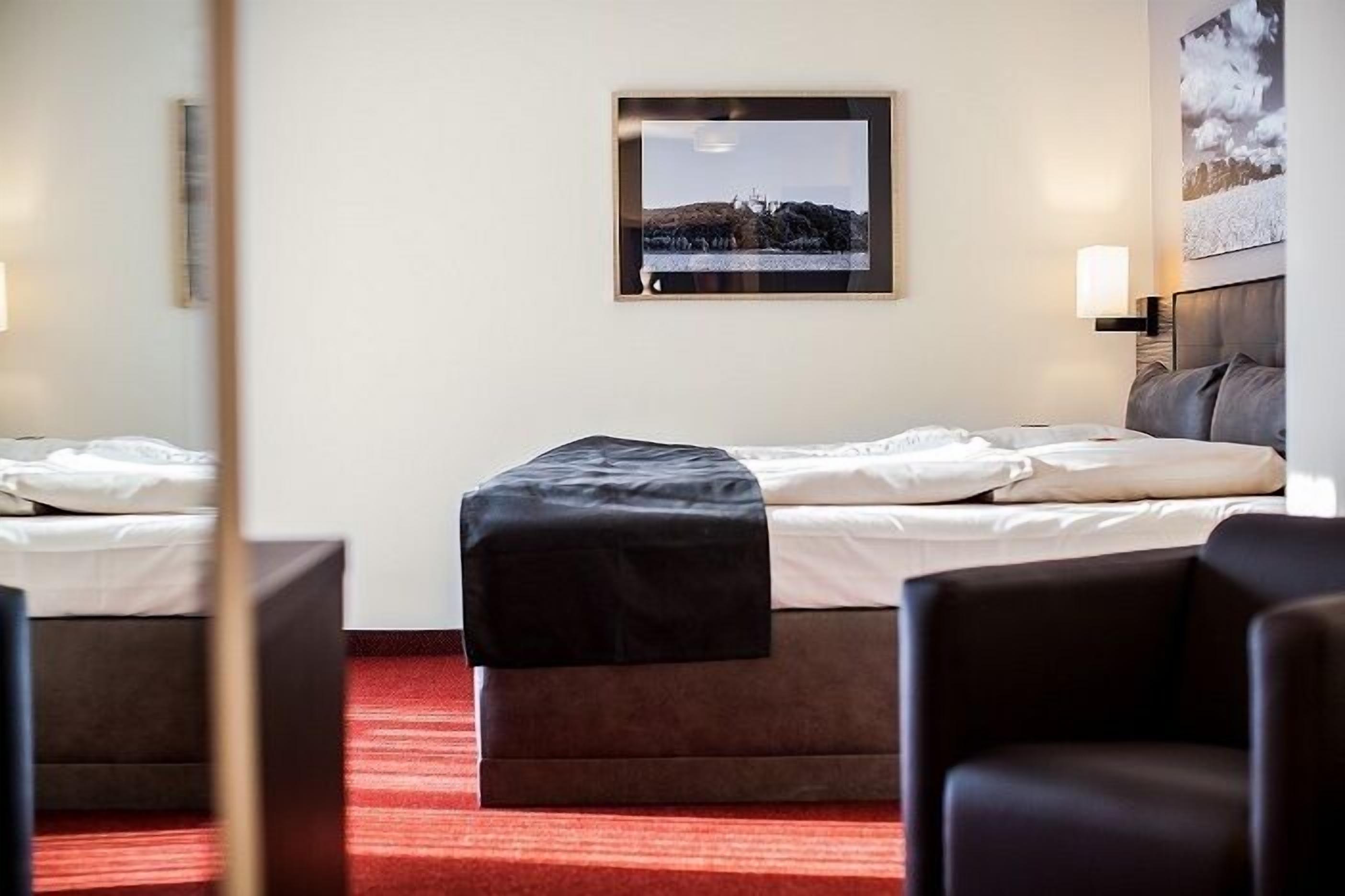 Une chambre d’hôtel avec un lit, une petite table, une chaise, un tableau au mur et une moquette rouge.