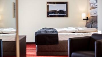 A hotel room with a bed, a small table, a chair, a picture on the wall, and a red carpet.