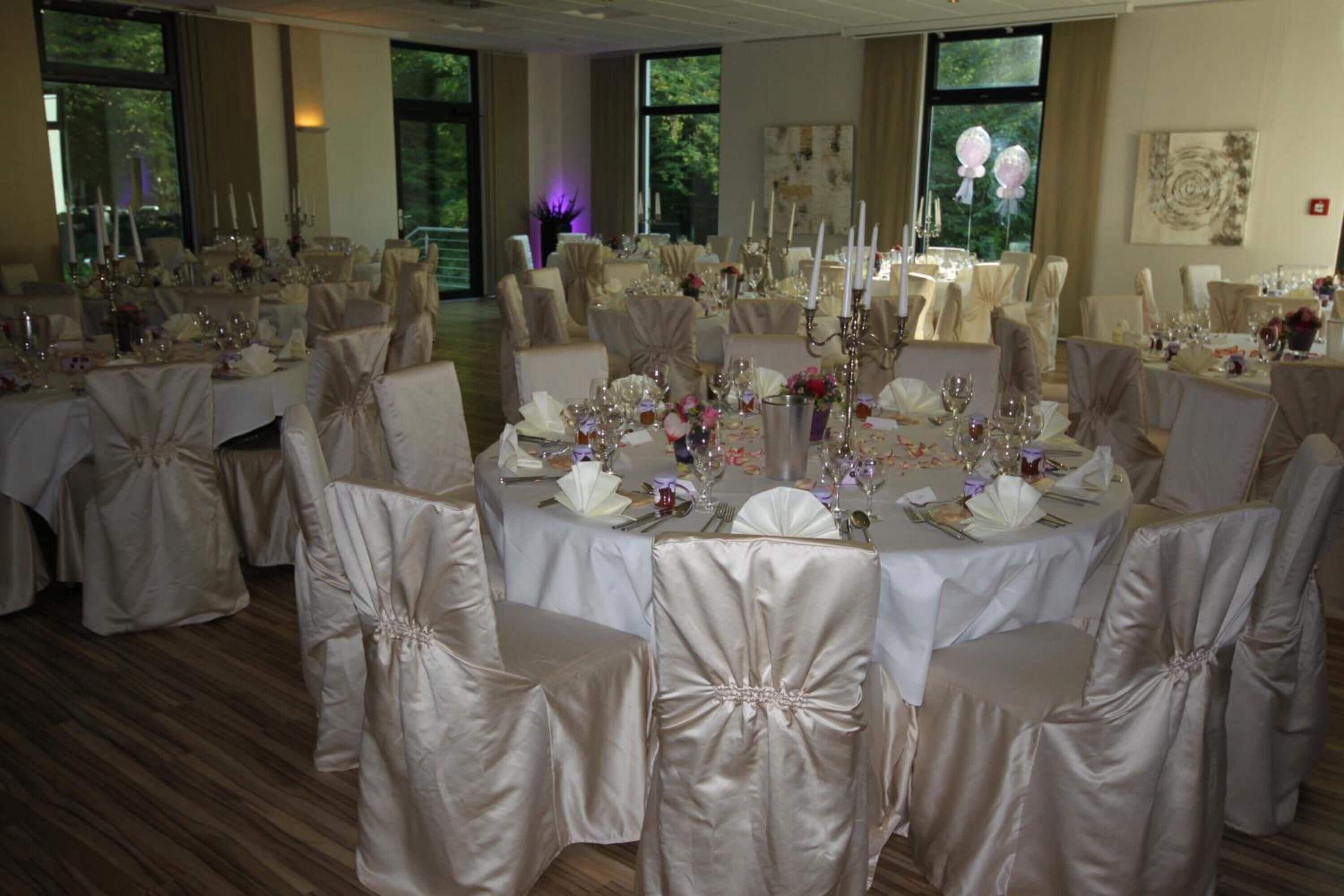 Uma sala de banquetes com mesas redondas preparadas para um evento formal, cada uma com toalhas brancas e cadeiras cobertas com tecido branco.