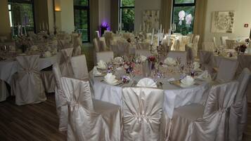 Uma sala de banquetes com mesas redondas preparadas para um evento formal, cada uma com toalhas brancas e cadeiras cobertas com tecido branco.