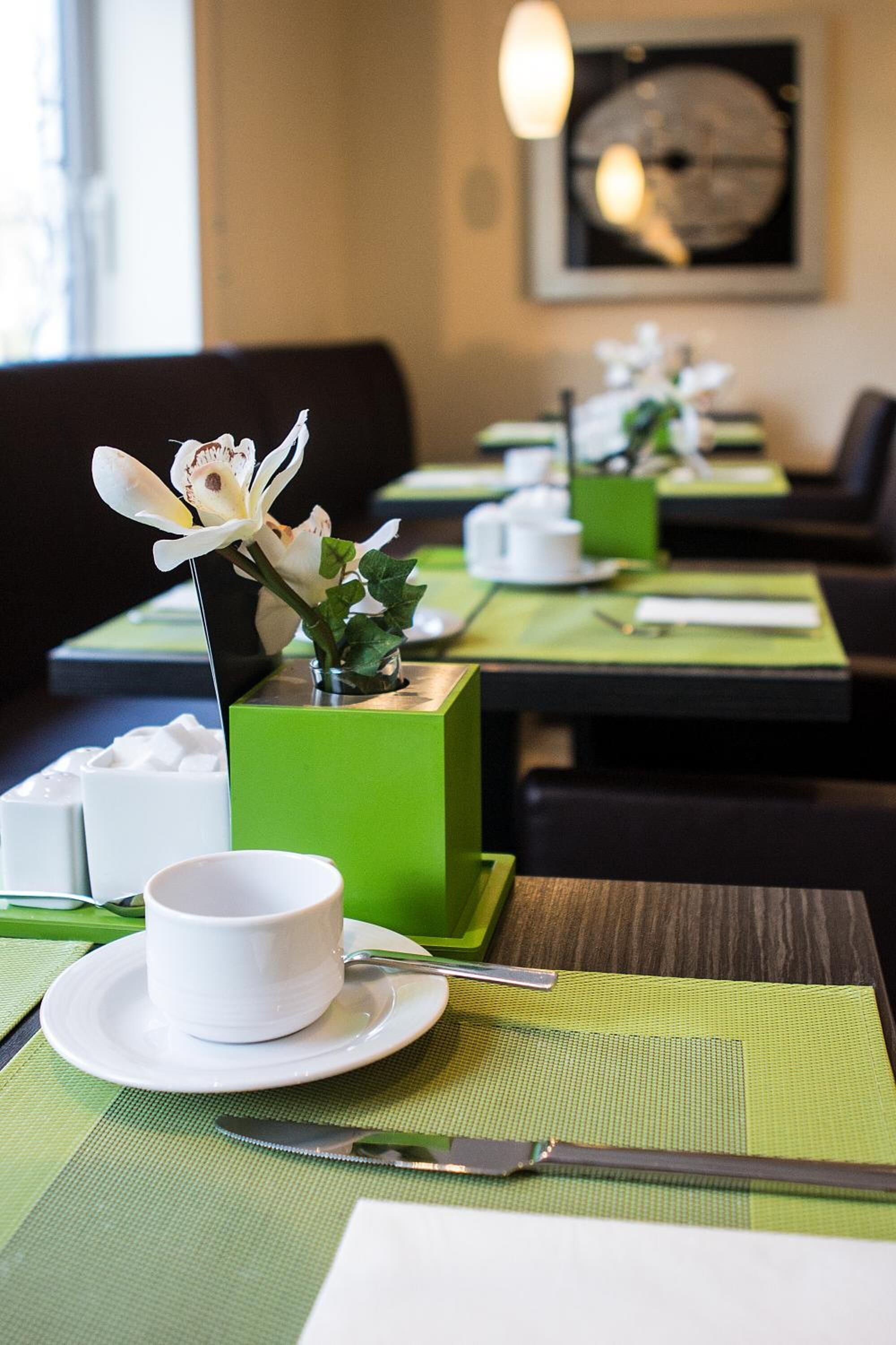 Un ambiente de restaurante con manteles individuales verdes, vajilla blanca y una sola orquídea decorando cada mesa.