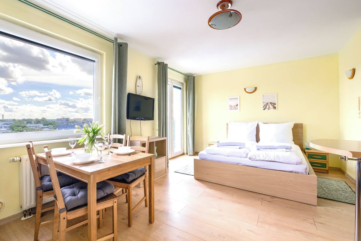 A room with a bed, a dining table with chairs, a TV, and a view of the city through a large window.