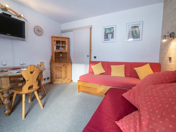 Een gezellige kamer met een rode bank, een houten eettafel, stoelen, een kast en een televisie aan de muur.