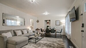 Ein modernes Wohnzimmer mit einem grauen Ecksofa, einem Glastisch und einem gemusterten Teppich. Es gibt eine Essecke mit Tisch und Stühlen, sowie einen an der Wand befestigten Fernseher.