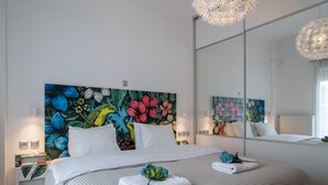 A modern hotel room with a large bed, white linens, a floral headboard, a large mirror, and a nightstand with a lamp.