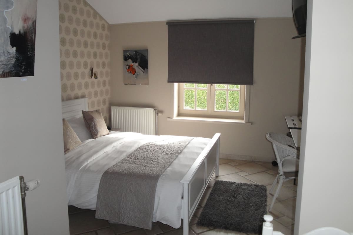 Une chambre à coucher avec un lit, un radiateur, une fenêtre avec un store, une chaise et un petit tapis.