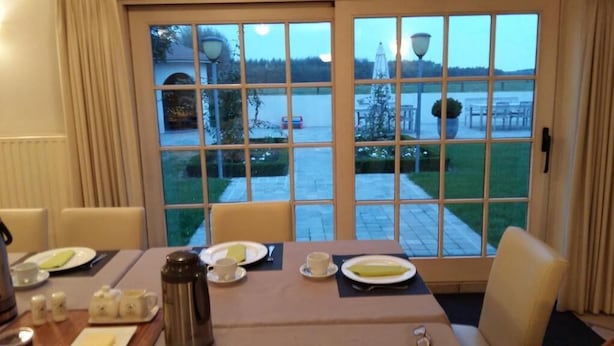 Comedor con una mesa dispuesta para dos personas, vista a un estacionamiento y zona de estar exterior a través de amplios ventanales.