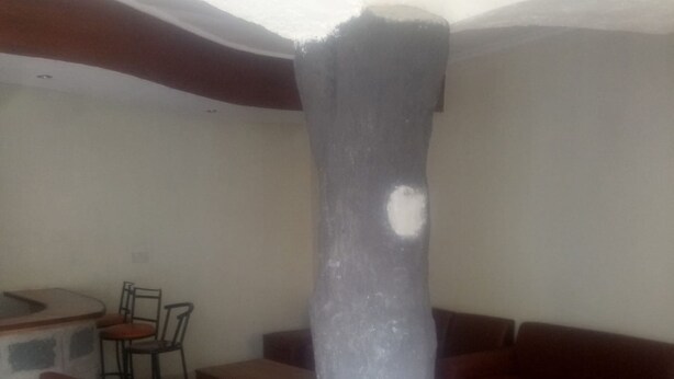 A room with a large, dark pillar, a red sofa, a wooden table, and a small bar area. - The Reeds Hotel (Ongata Rongai)