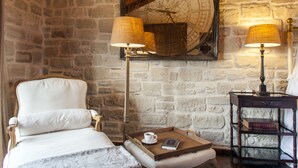 Een kamer met een stenen muur, een bed met een wit hoofdeinde, twee lampen, een dienblad met een kopje en een boek, en een houten nachtkastje.