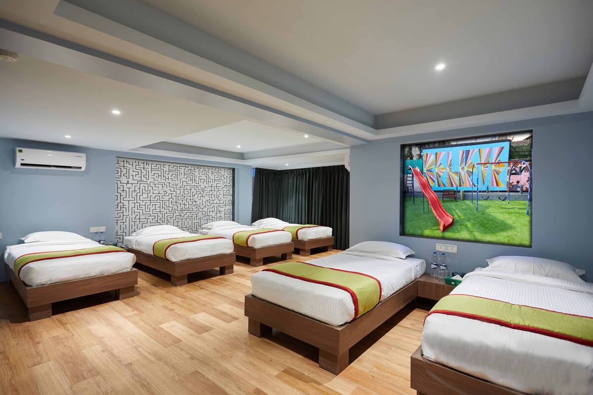 A hotel room with four beds arranged in a U-shape, a large window with a view of a playground, and a wall-mounted air conditioning unit.