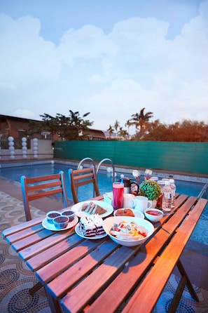 Una mesa de madera con diversos alimentos y bebidas junto a una piscina, con vistas claras a la piscina y palmeras al fondo.