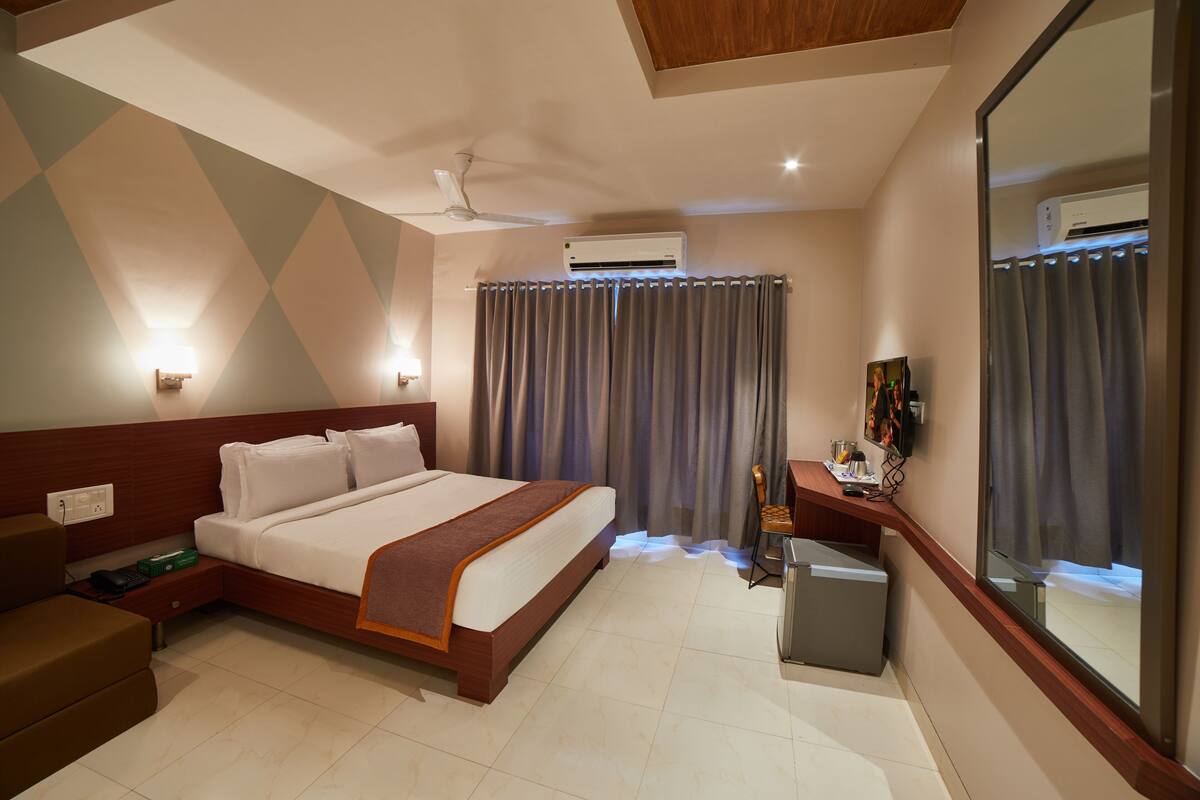 A hotel room with a large bed, a desk with a chair, a small table, an air conditioning unit, and a mirror.