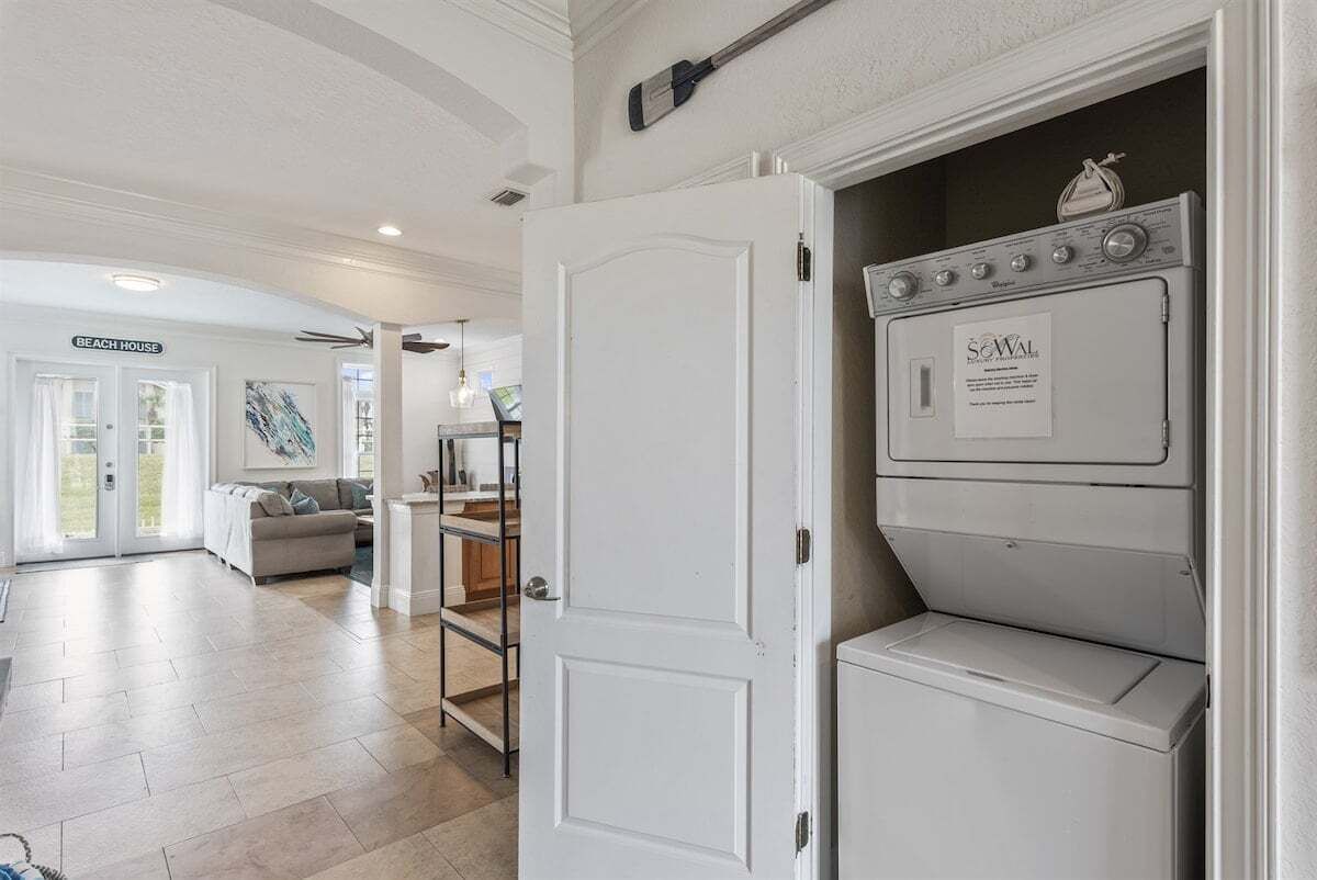 A laundry room with a front-loading washer and dryer, a door leading to a living area, and a sign that reads 'BEACH HOUSE'.