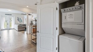 Un lavadero con lavadora y secadora de carga frontal, una puerta que da a una sala de estar, y un letrero que dice 'BEACH HOUSE'.
