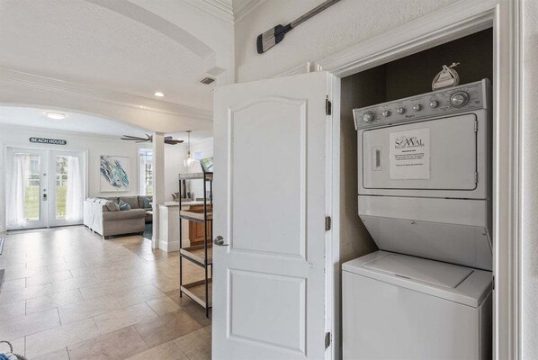 A laundry room with a front-loading washer and dryer, a door leading to a living area, and a sign that reads 'BEACH HOUSE'.