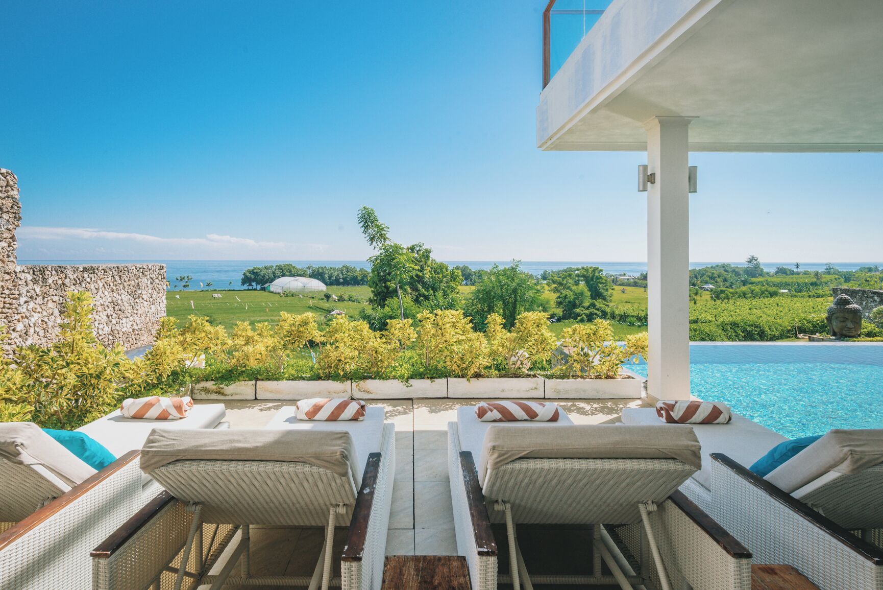 A poolside lounge with wicker chairs and a view of the ocean and greenery.