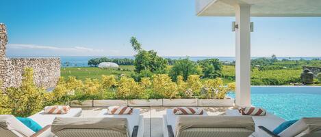 A poolside lounge with wicker chairs and a view of the ocean and greenery.