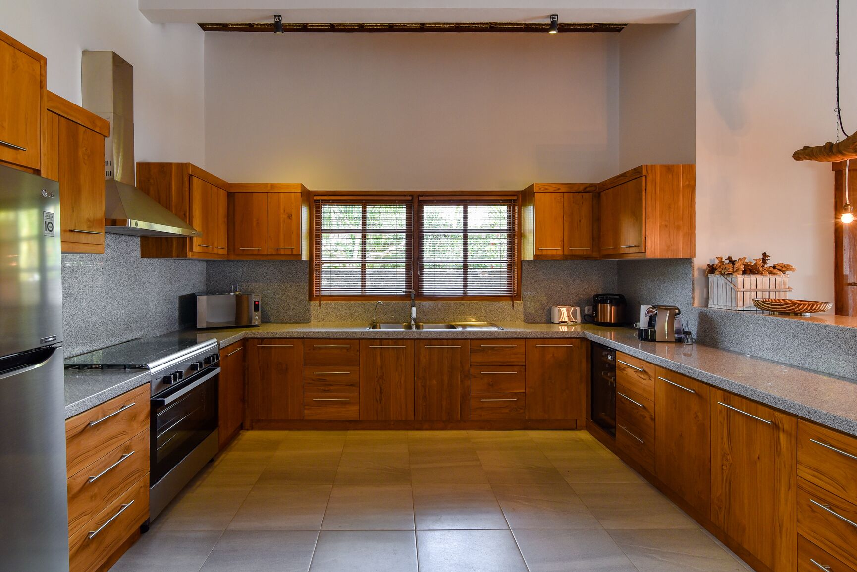 A modern kitchen with wooden cabinets, granite countertops, a stainless steel refrigerator, a built-in oven, and a window with blinds.