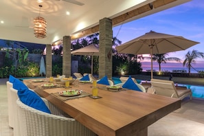 A dining area with a wooden table, blue cushions, and umbrellas, overlooking a pool and beach at sunset. - Villa Saneva (Seririt)