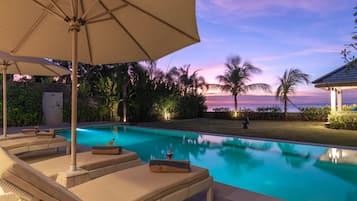Zona de piscina con sillones de descanso y una sombrilla, rodeada de vegetación y vistas al océano al atardecer.