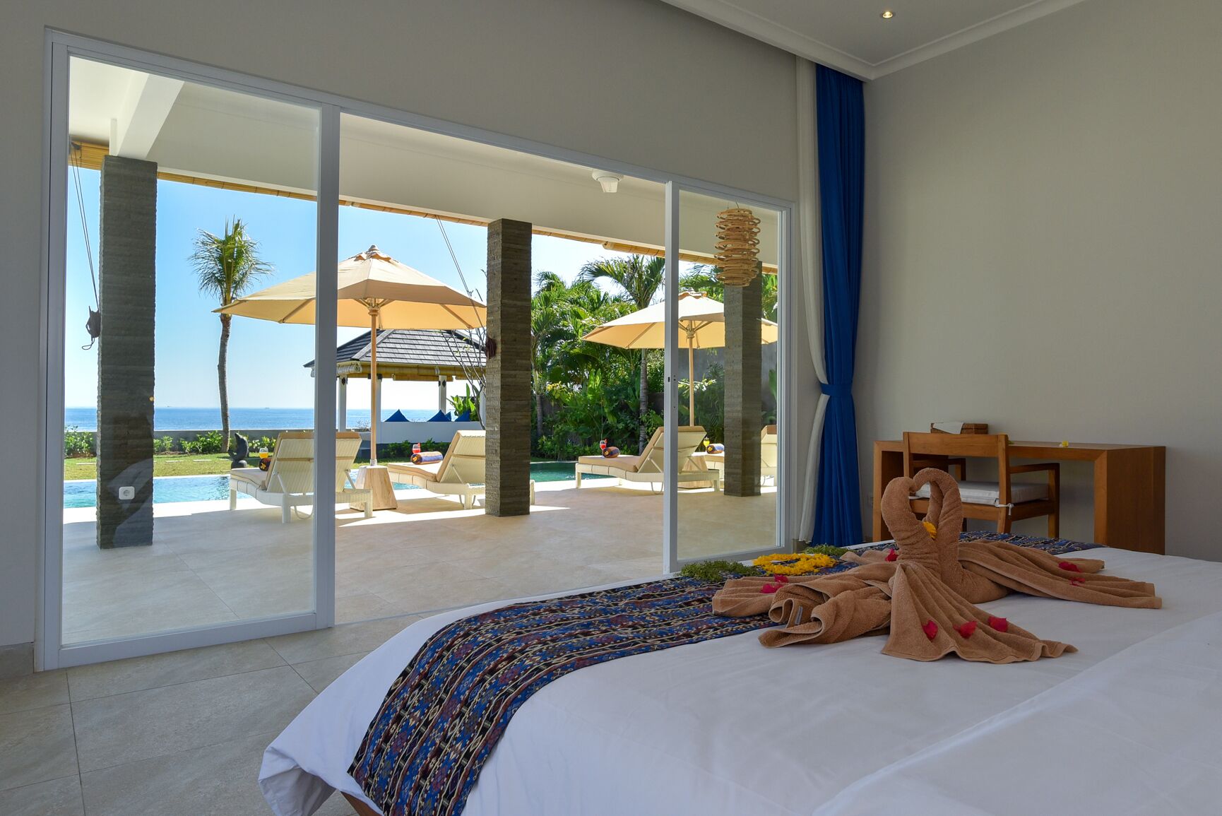 Habitación de hotel con una cama grande, vistas al océano y un patio con sillas de descanso y sombrillas.