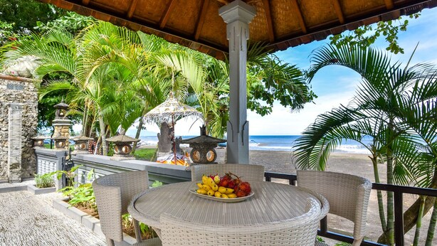 Ein überdachter Außenbereich mit Korbmöbeln und einem Tisch, der mit Obst bedeckt ist, mit Blick auf einen Strand.