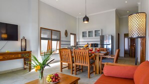 Ein modernes Wohnzimmer mit Essbereich, einer Küche im Hintergrund und einem großen Fenster mit Jalousien.