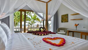 Ein Zimmer mit einem Bett, einer blumigen Herz-Arrangement und Blick auf Palmen und den Strand.