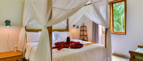 A room with a four-poster bed, a wooden nightstand, a small table, and a window with a view of greenery.
