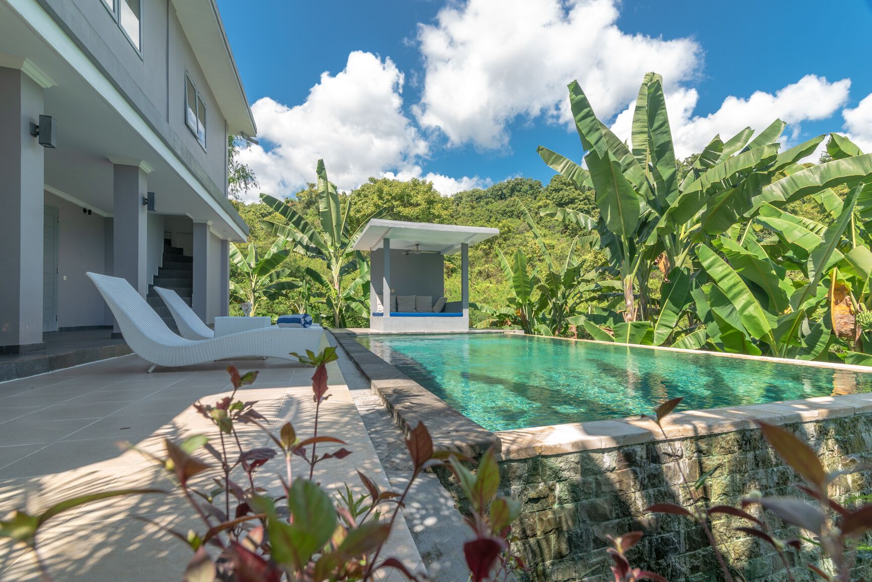A modern pool area with lounge chairs, a small pavilion, and lush tropical vegetation.
