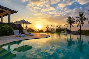 Una piscina con sillas de descanso y una sombrilla, rodeada de palmeras y un atardecer.