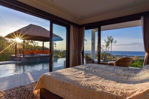 Une chambre avec un grand lit, une vue sur une piscine et un espace extérieur, et un coucher de soleil.